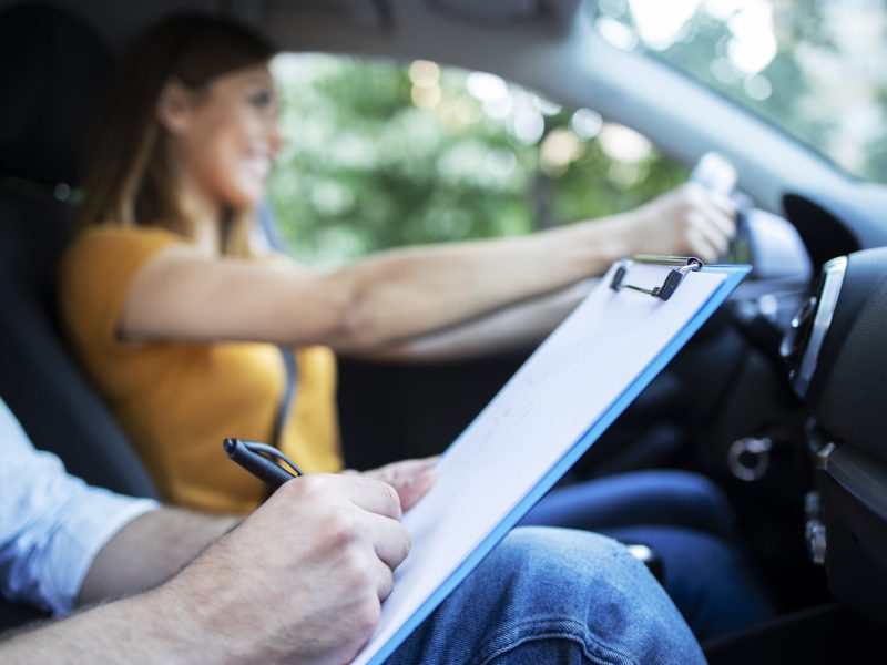 Close up view of driving instructor holding checklist while in background female student steering and driving car.